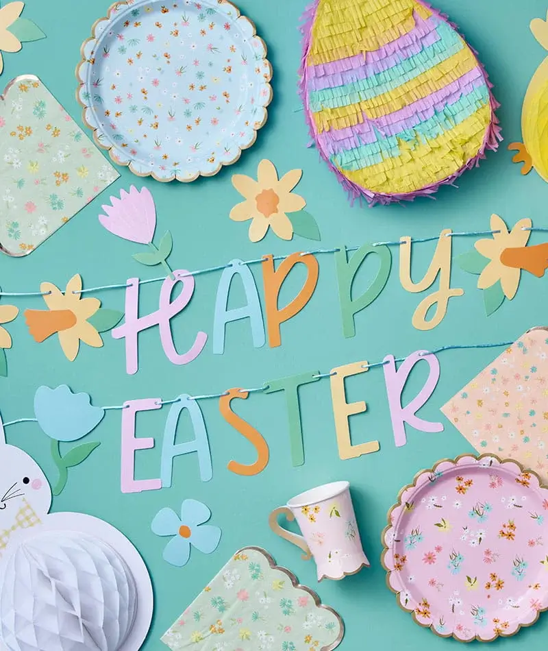 Colourful Easter decorations arranged on a teal background, featuring a Happy Easter banner, floral cut-outs, paper cups and a yellow chick decoration.